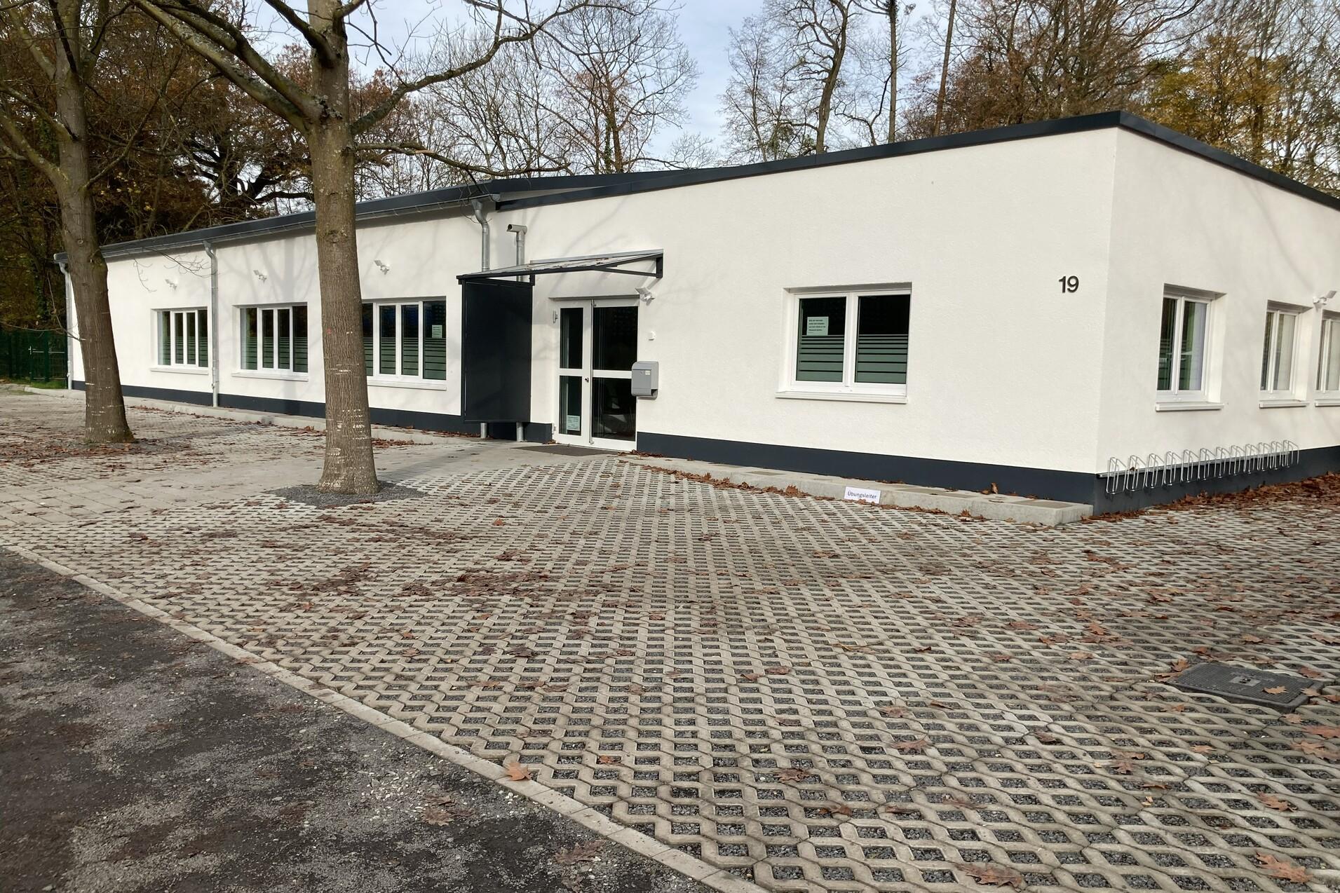 Moderne weiße Gebäudeansicht mit großen Fenstern und gepflastertem Vorplatz, umgeben von Bäumen.