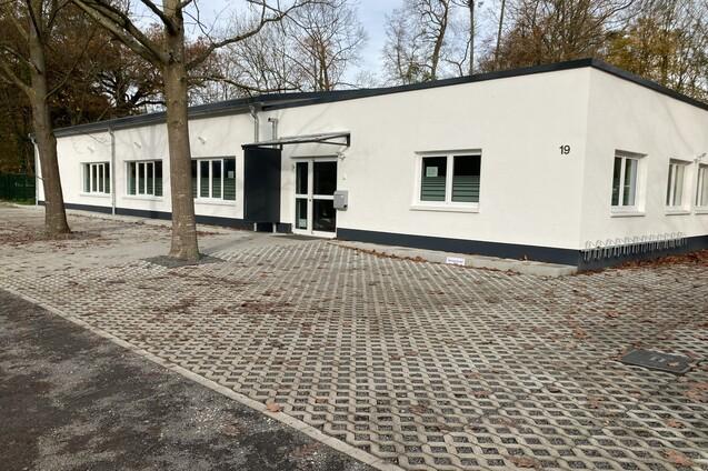 Moderne wei&szlig;e Geb&auml;udeansicht mit gro&szlig;en Fenstern und gepflastertem Vorplatz, umgeben von B&auml;umen.