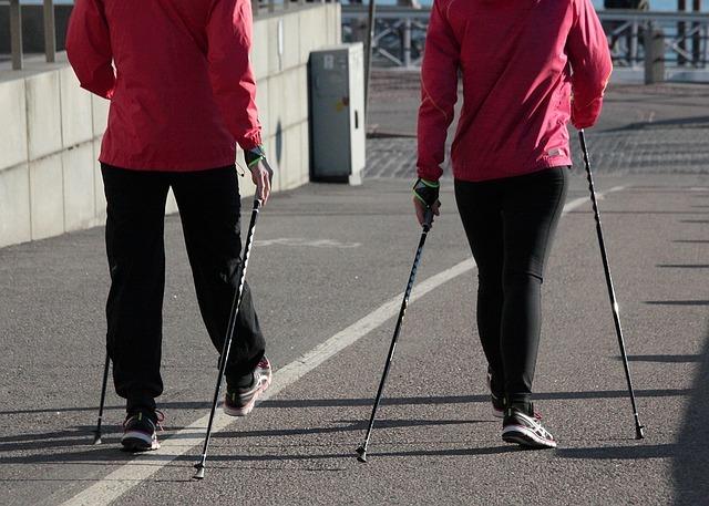 Zwei Personen in Rosa tragen Nordic-Walking-Stöcke und gehen auf einem Gehweg entlang einer Uferpromenade.