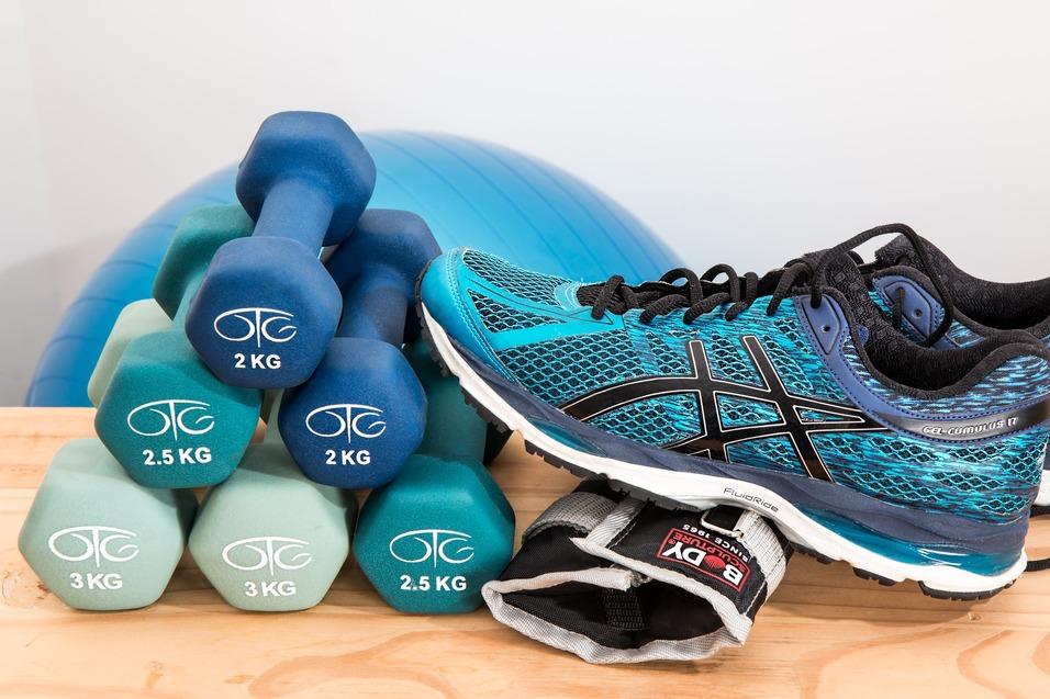 Sportliches Setup mit Handgewichten in verschiedenen Größen und einem blauen Laufschuh auf einer Holzoberfläche.