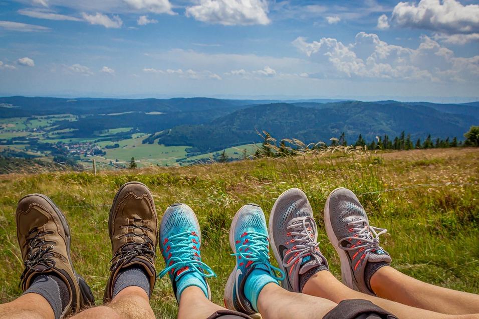 F&uuml;&szlig;e in Wanderschuhen und Sportschuhen stehen auf einer Wiese mit Aussicht auf eine gr&uuml;ne Berglandschaft.