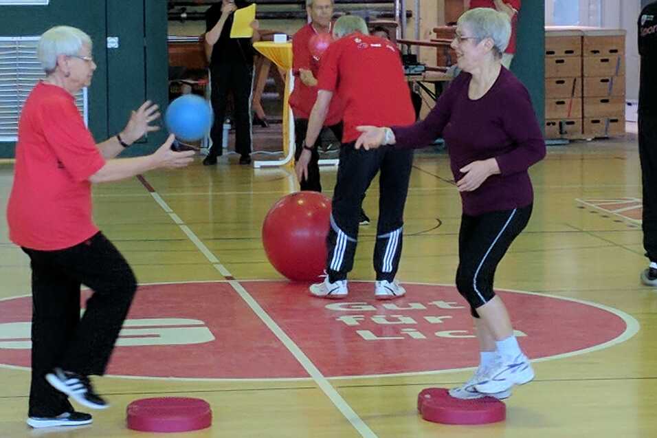 Zwei ältere Frauen balancieren auf Podesten und werfen sich einen blauen Ball in einer Sporthalle zu.