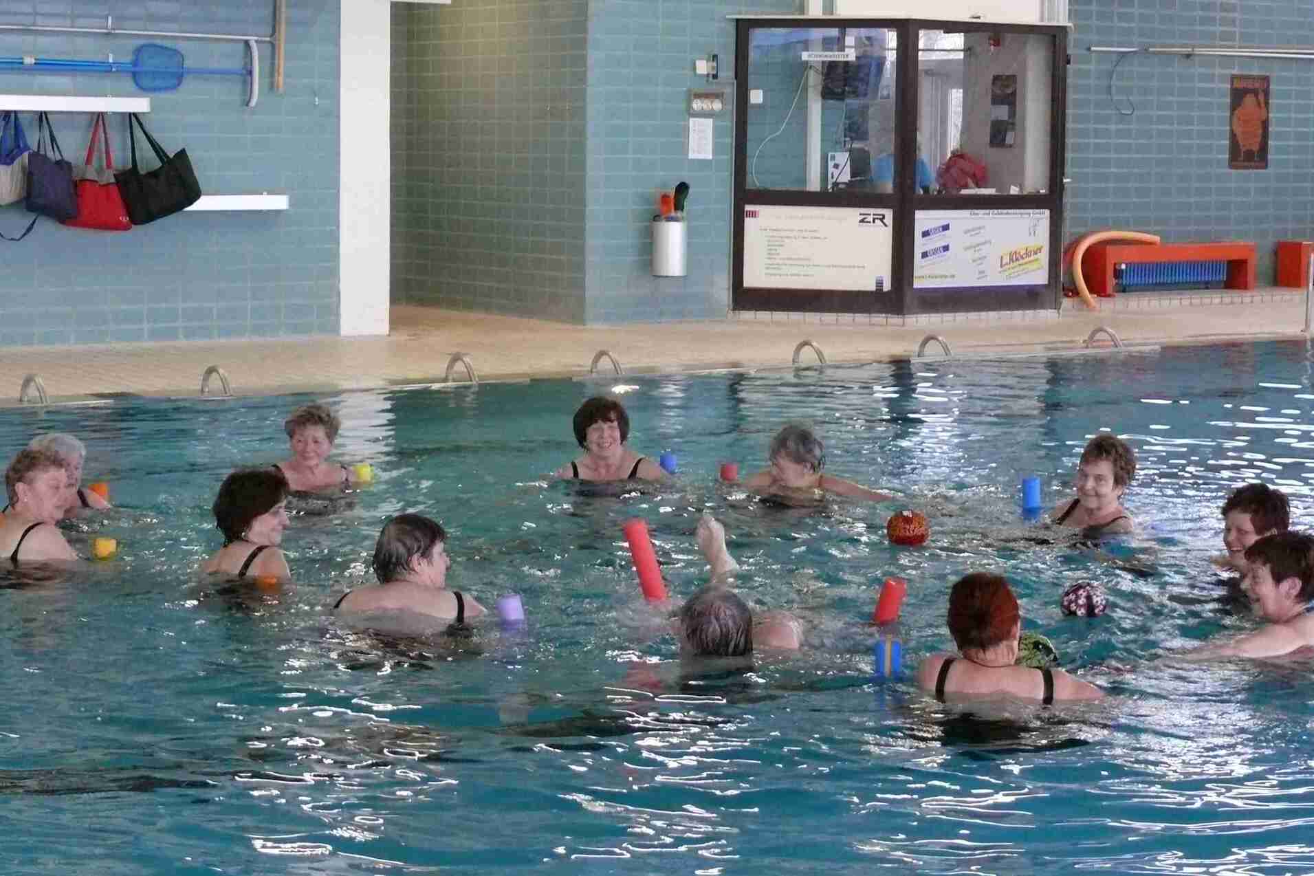 Gruppentraining im Wasser: Frauen mit Schwimmnudeln und Bällen stehen in einem Schwimmbecken.
