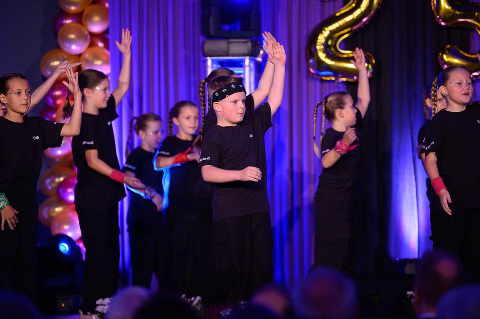 Gruppe von Kindern in schwarzen Outfits, die auf einer Bühne tanzen, beleuchtet von bunten Lichtern und Luftballons.