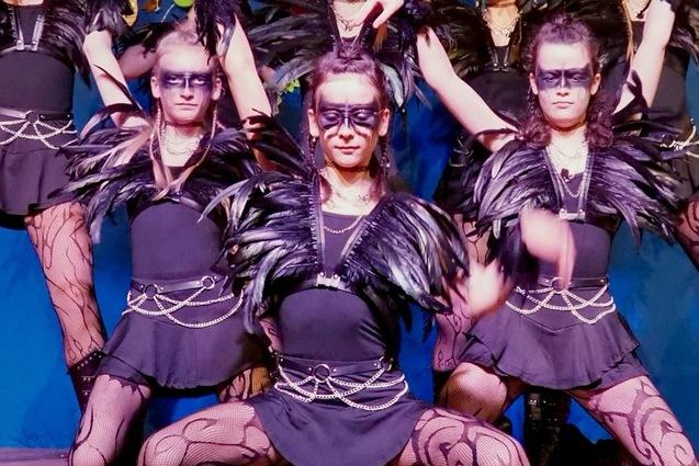 Tanzgruppe in schwarz gekleideten Kost&uuml;men mit Federn und Masken, die auf einer B&uuml;hne vor bunten Luftballons auftritt.