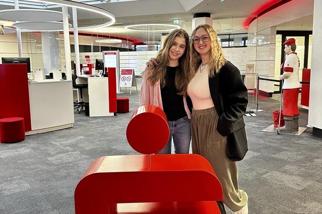 Zwei junge Frauen stehen l&auml;chelnd neben einem roten S-Logo und einem F&ouml;rderungsschild in einer modernen Bankfiliale.