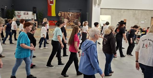 Gruppe von Erwachsenen in verschiedenen Shirts, die synchron in einer hellen Halle tanzen. Im Hintergrund eine Dekoration.