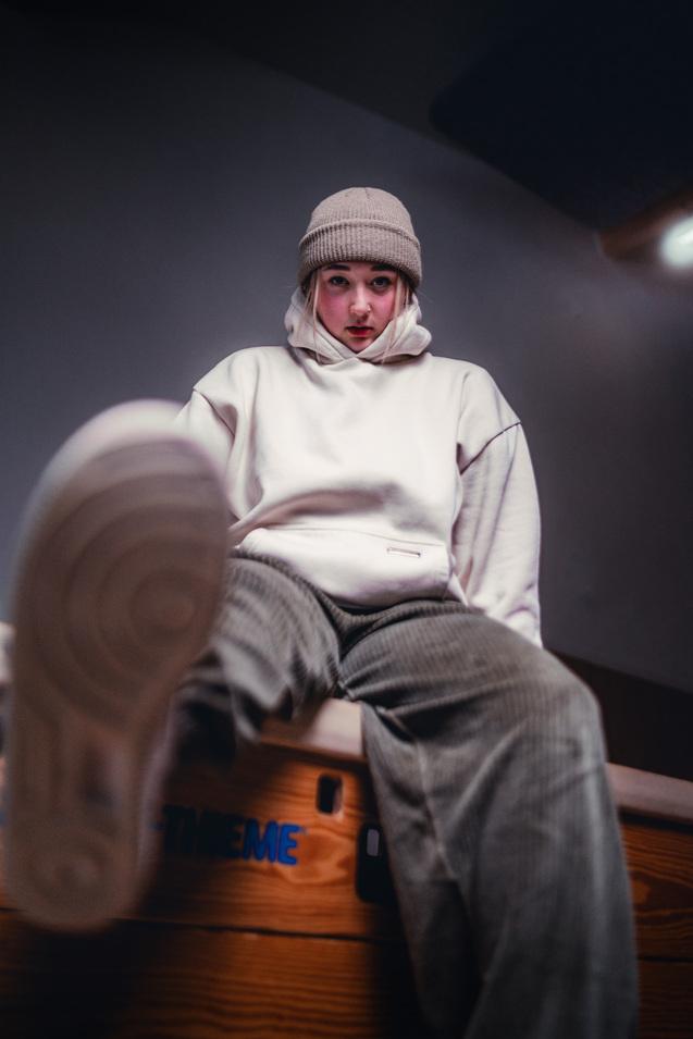 Jugendliche Person in beiger M&uuml;tze und wei&szlig;em Hoodie sitzt auf einer Holzkante und richtet den Blick in die Kamera.
