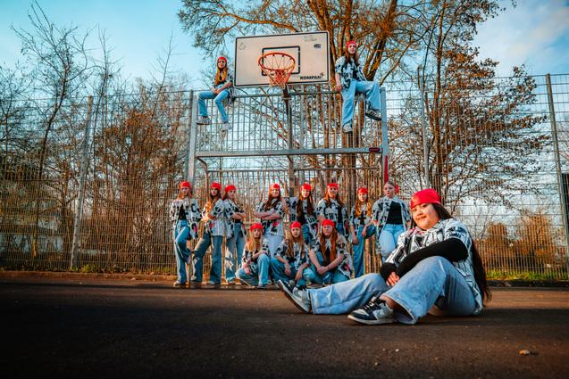 Gruppe von zw&ouml;lf Personen in roten M&uuml;tzen auf einem Basketballplatz, einige sitzen, andere stehen am Korb.