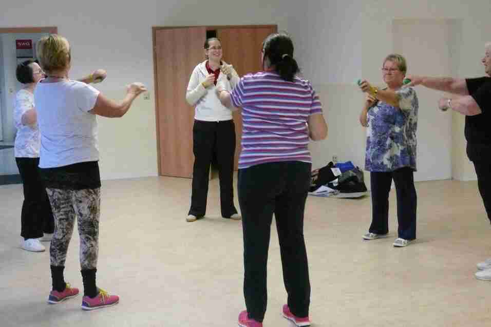Gruppe von Frauen macht in einem hellen Raum gemeinsam Bewegungs&uuml;bungen unter Anleitung einer Trainerin.