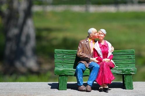 Figuren eines &auml;lteren Paares sitzen eng umschlungen auf einer gr&uuml;nen Bank im Freien vor unscharfem Hintergrund.