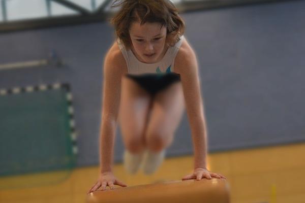Kind springt vom Sprungbrett ab, H&auml;nde auf dem Ger&auml;t, in einer Sporthalle mit bunten W&auml;nden im Hintergrund.