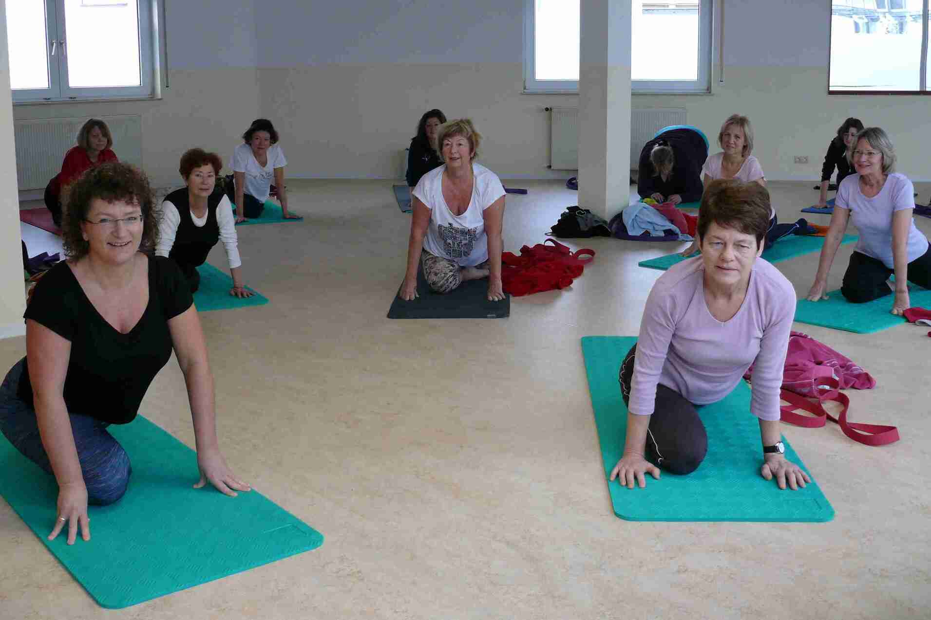 Gruppe von Frauen beim Yoga auf Matte, verschiedene Übungen in einem hellen Raum mit Fenstern.