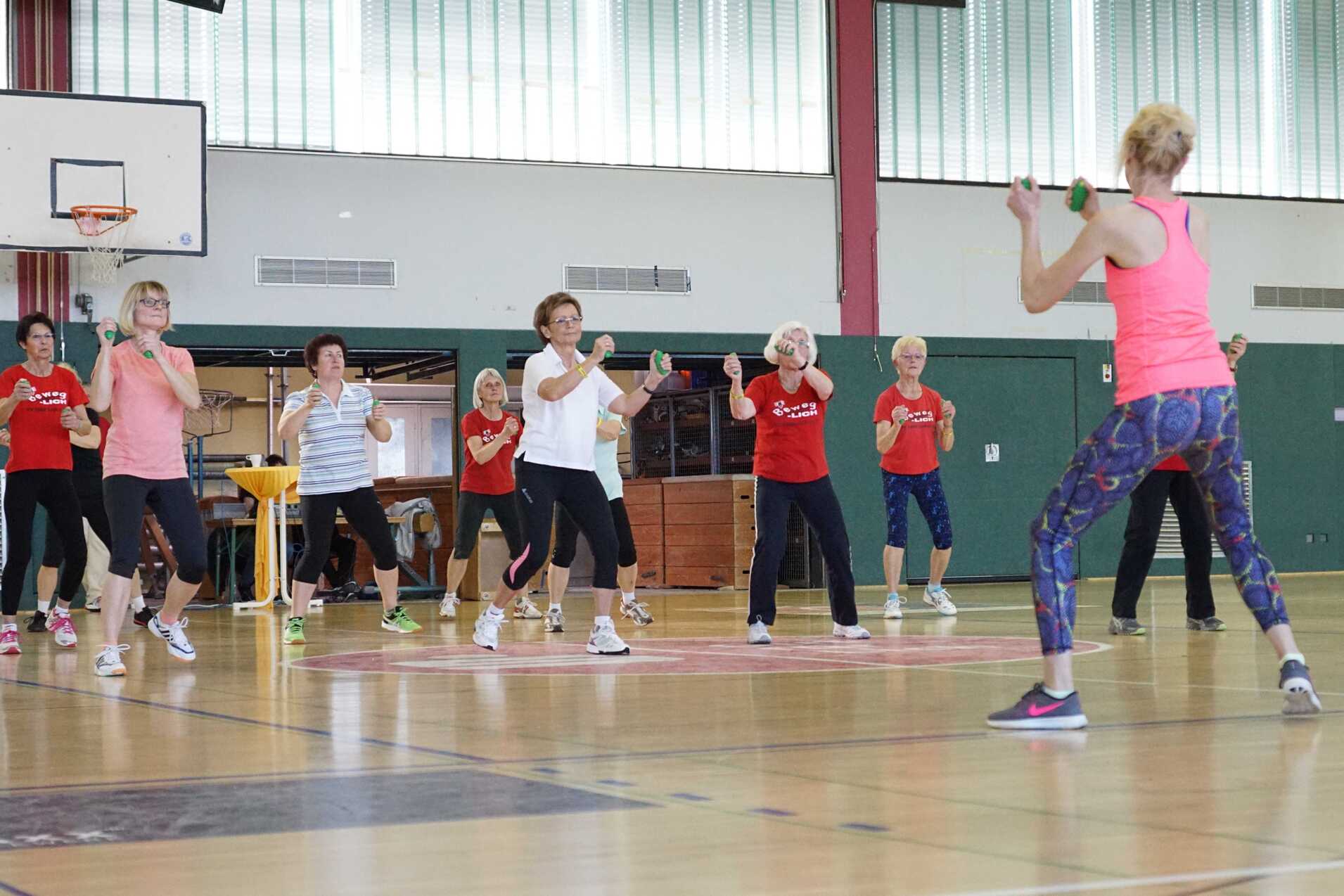 Gruppe älterer Frauen beim Fitness-Training in einer Sporthalle, angeleitet von einer Trainerin mit Hanteln.