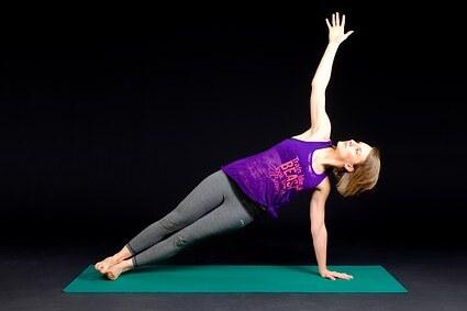 Frau in seitlicher Planke auf einer Yoga-Matte, eine Hand nach oben ausgestreckt, vor schwarzem Hintergrund.