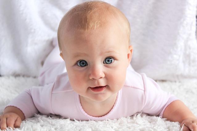 Ein Baby liegt auf dem Bauch auf einer weichen Decke und blickt mit großen, blauen Augen direkt in die Kamera.