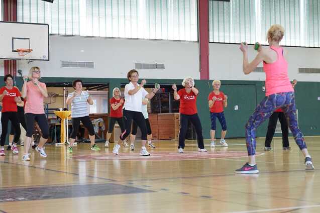 Gruppe &auml;lterer Frauen beim Fitness-Training in einer Sporthalle, angeleitet von einer Trainerin mit Hanteln.