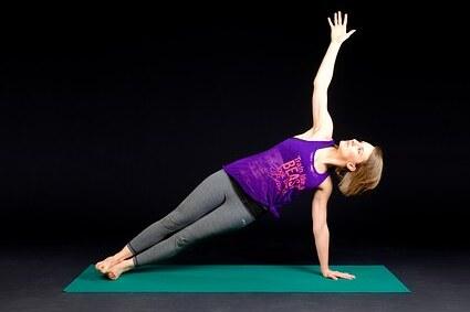 Frau in seitlicher Planke auf einer Yoga-Matte, eine Hand nach oben ausgestreckt, vor schwarzem Hintergrund.