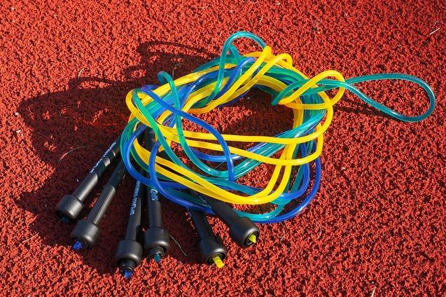 Bunte Springseile mit schwarzen Griffen liegen auf einer roten Sportplatzoberfl&auml;che.