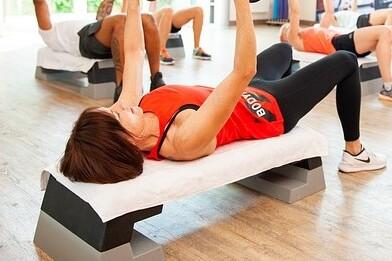 Frau in rotem Shirt hebt Gewichte auf einem Trainingsger&auml;t in einem hellen Fitnessstudio. Weitere Personen im Hintergrund.