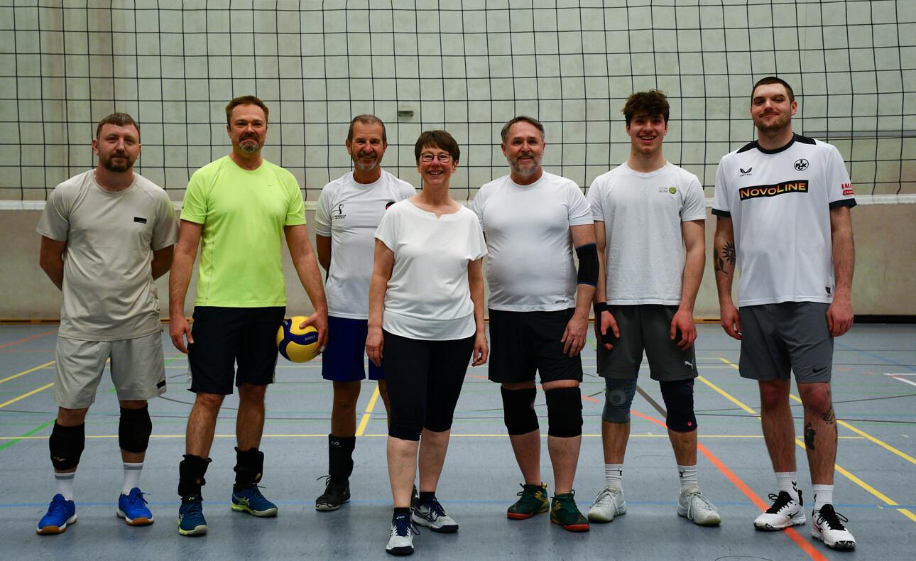 Gruppe von acht Personen in Sportkleidung stehen vor einem Volleyballnetz in einer Halle. Ein Spieler hält einen Volleyball.