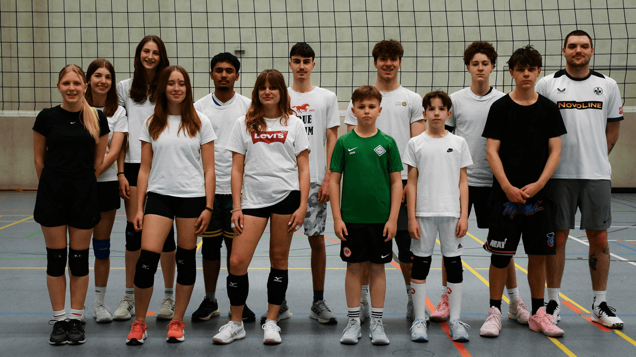 Gruppe von 15 Volleyballspielern in Sportkleidung auf einem Hallenboden, neben einem Volleyballnetz aufgestellt.