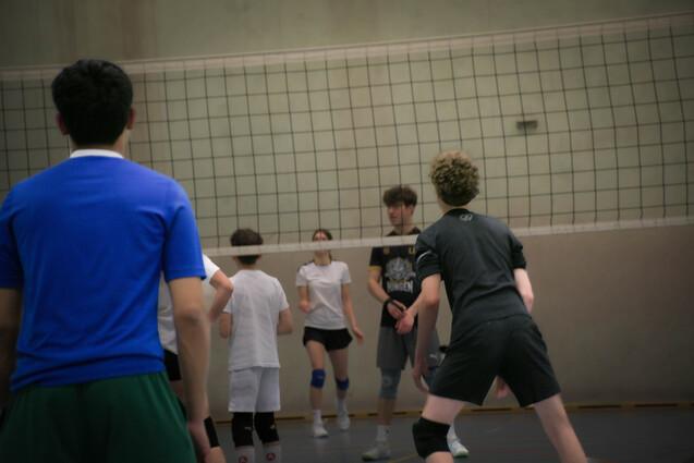 Gruppe von Jugendlichen spielt Volleyball in einer Halle, Netz im Vordergrund, Spieler in unterschiedlichen Trikots.