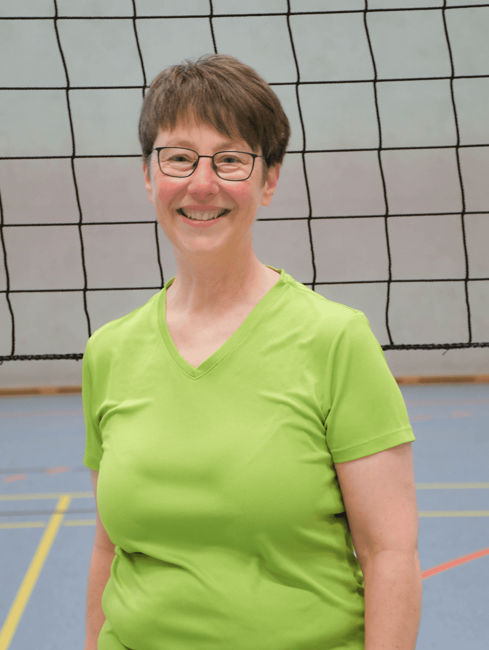 Frau mit Brille trägt ein grünes Sportshirt und steht vor einem Volleyballnetz in einer Sporthalle.