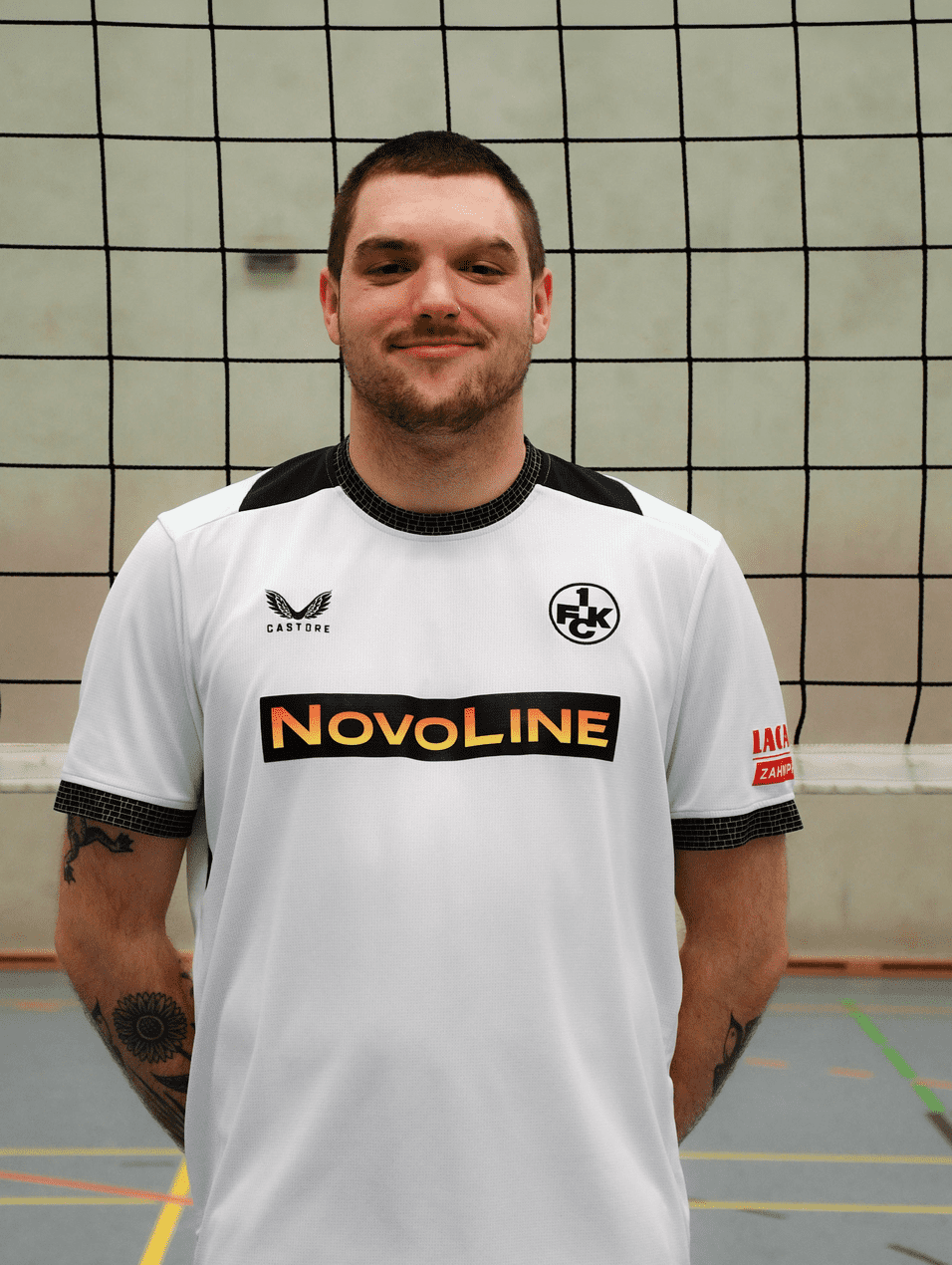 Mann in weißem Sporttrikot mit Sponsorenlogo steht vor einem Volleyballnetz in einer Sporthalle.