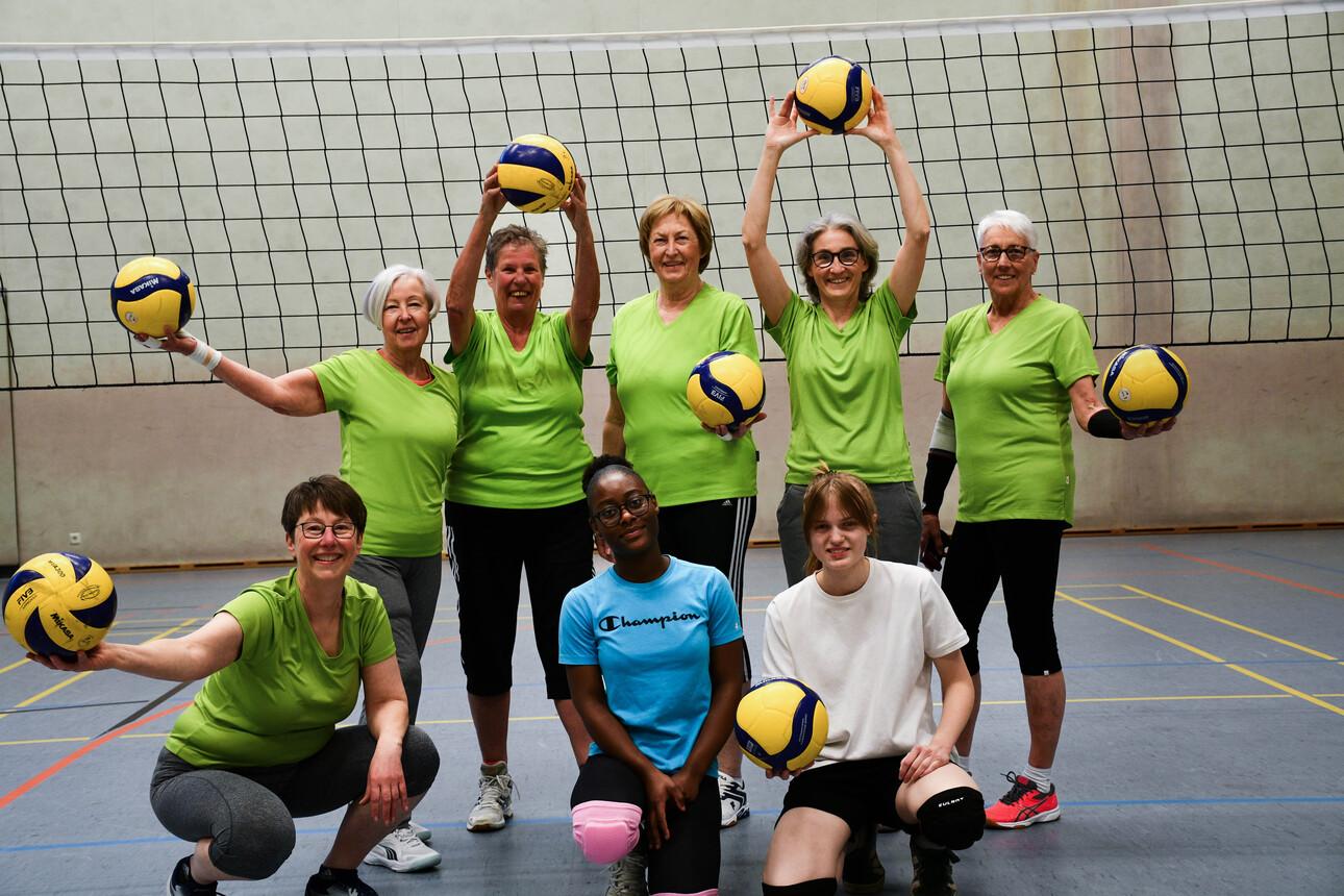 Gruppe von acht Frauen in grünen T-Shirts, die Volleyballbälle halten, vor einem Volleyballnetz in einer Sporthalle.