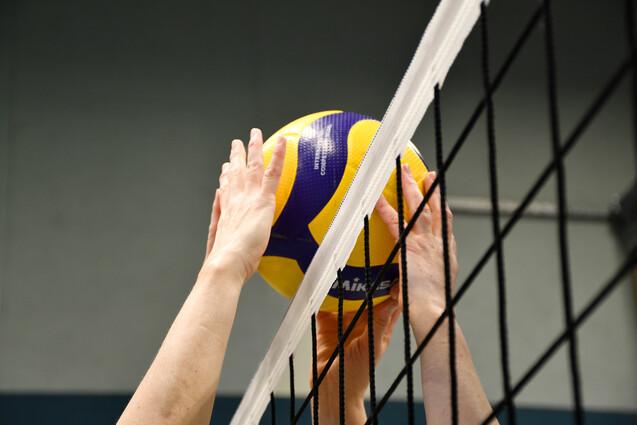 Zwei Hände strecken sich nach einem gelben und lila Volleyball über das Netz in einer Sporthalle.