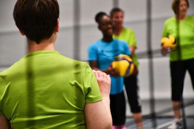 Vierschlägige Volleyballspielerinnen in Freizeitkleidung stehen auf dem Spielfeld und halten Bälle in den Händen.