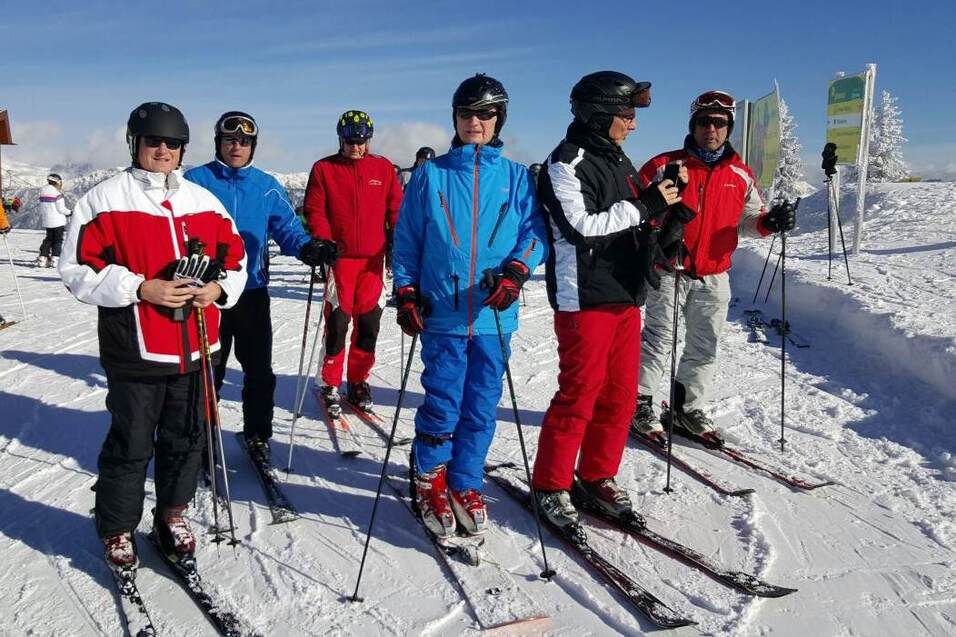 Sechs Skifahrer in bunten Outfits stehen auf einer verschneiten Piste unter blauem Himmel.