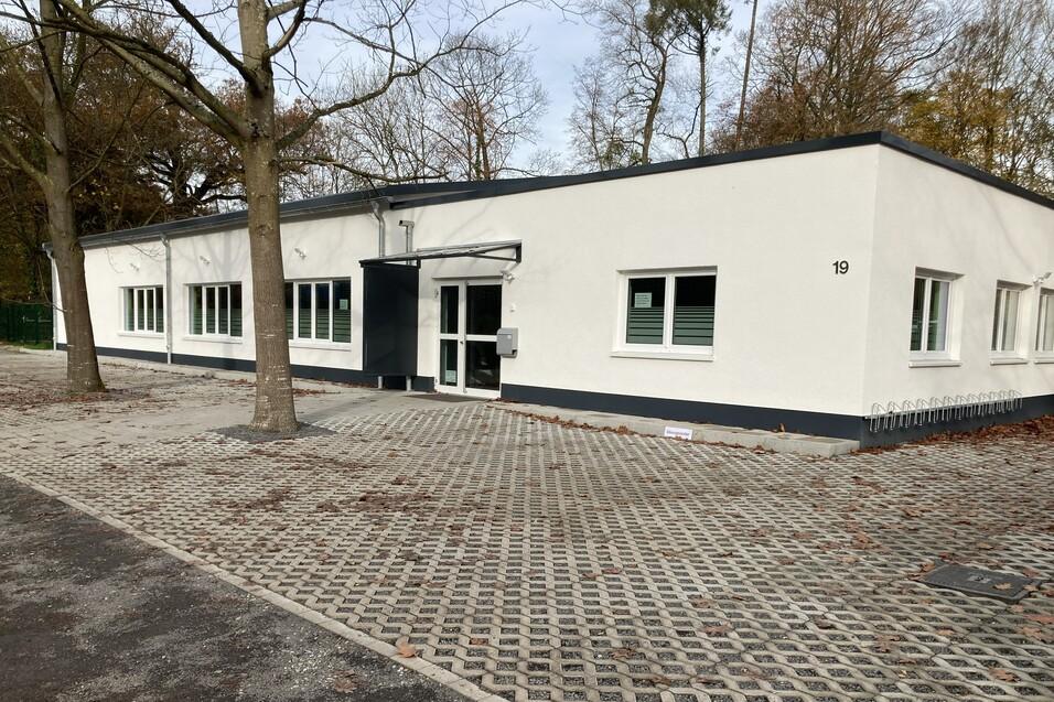 Moderne, einstöckige, weiße Gebäudefront mit Glasfenstern und einem Eingangsbereich, umgeben von einem gepflasterten Platz.