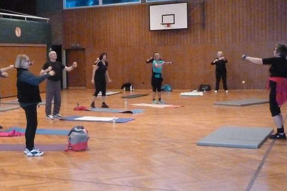 Eine Gruppe von sieben Personen macht Fitnessübungen in einer Sporthalle mit Holzfußboden und einem Basketballkorb.