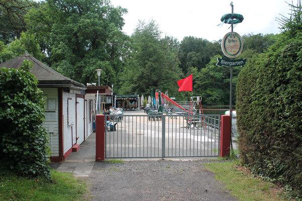 Eingang zum Biergarten mit Metallzaun, spielenden Kindern und einem roten Signalflaggen am Seeufer.