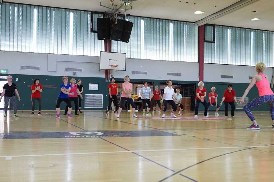 Gruppentraining in einer Sporthalle mit mehreren Teilnehmern, die einer Trainerin vor ihnen folgen.