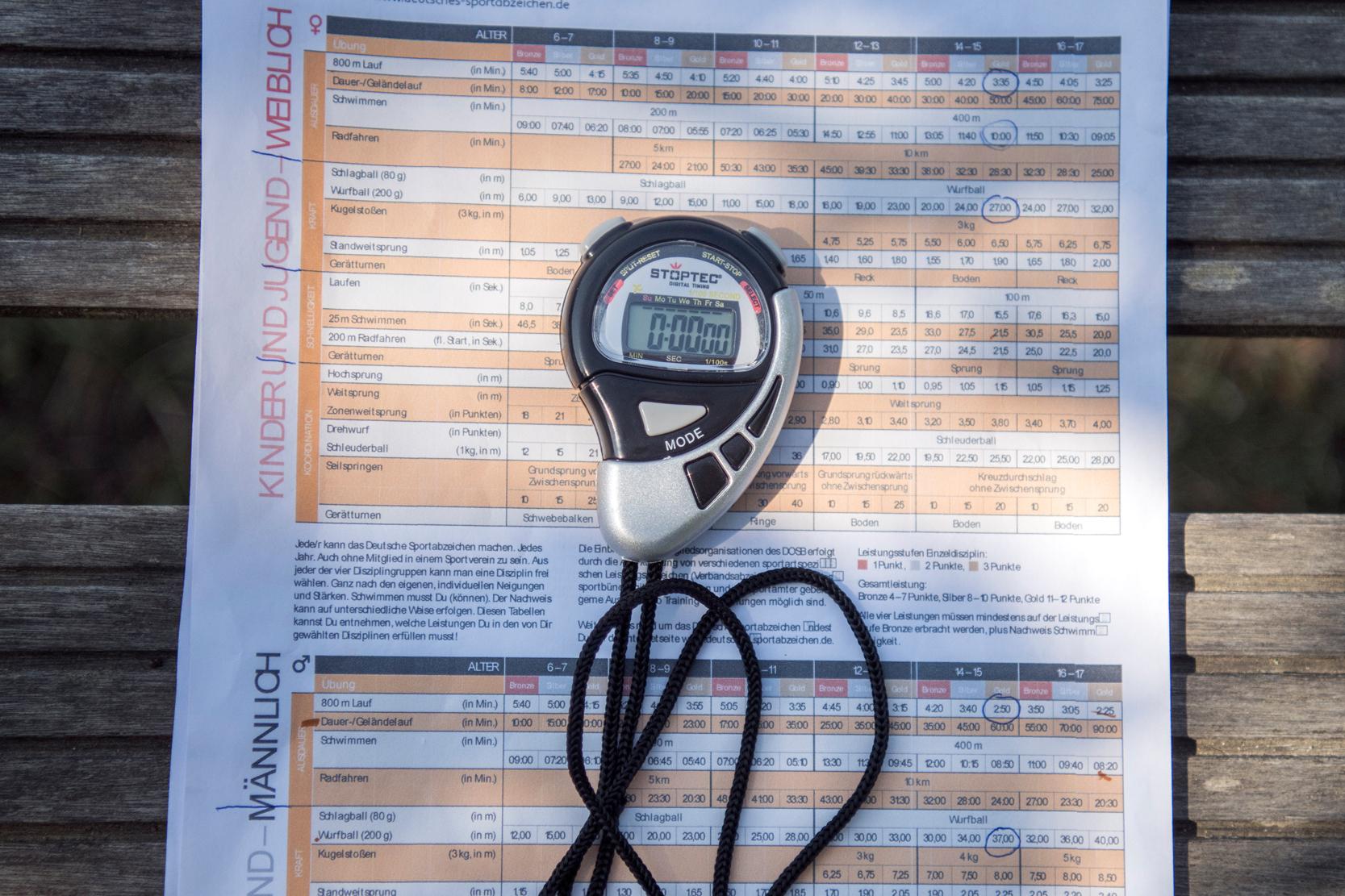 Stoppuhr und Infoblatt des Deutschen Sportabzeichens auf einer Holzbank im Freien.