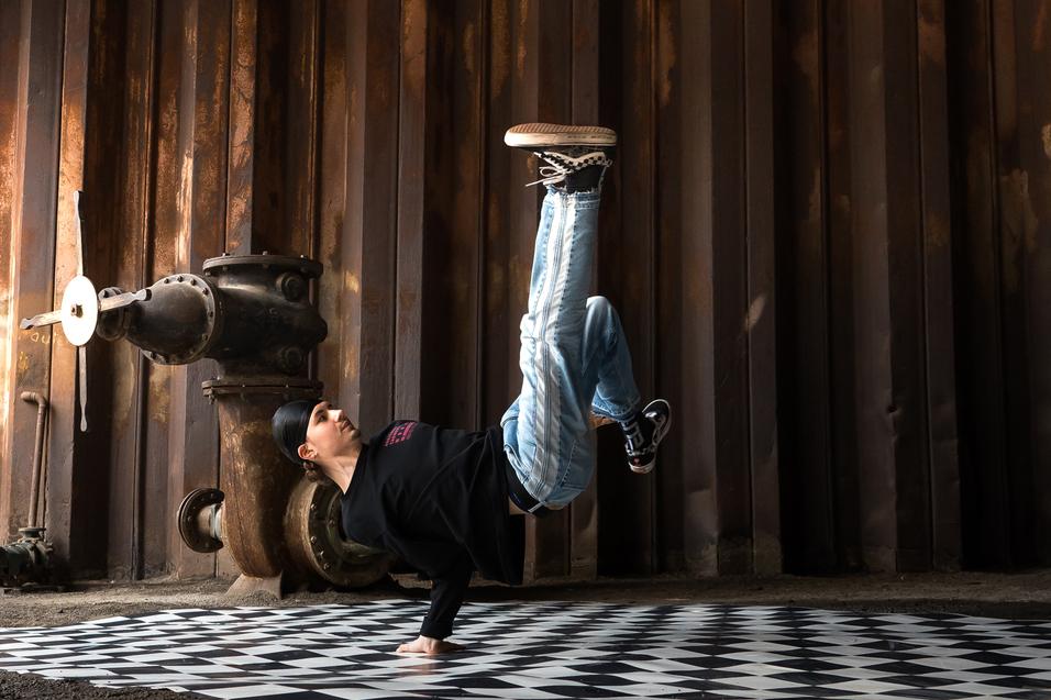 Junge in sportlicher Kleidung führt einen Handstand auf einem schachbrettartigen Boden in einer industriellen Umgebung aus.