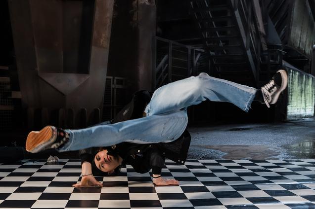 Tänzer führt einen Breakdance-Move auf einem schwarz-weißen Schachbrettboden durch, in einer modernen Umgebung.