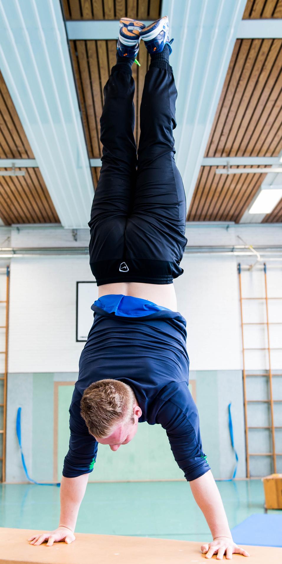 Sportler führt einen Handstand auf einer Kiste in einer Turnhalle durch, im Hintergrund sind Turngeräte sichtbar.